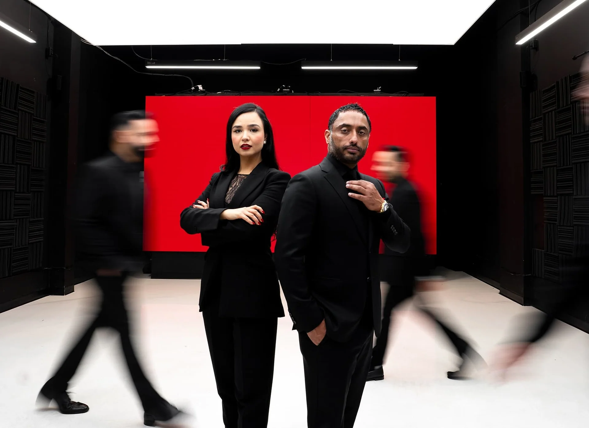 Two people in black standing still in front of a red screen in a studio while other people walk past blurred