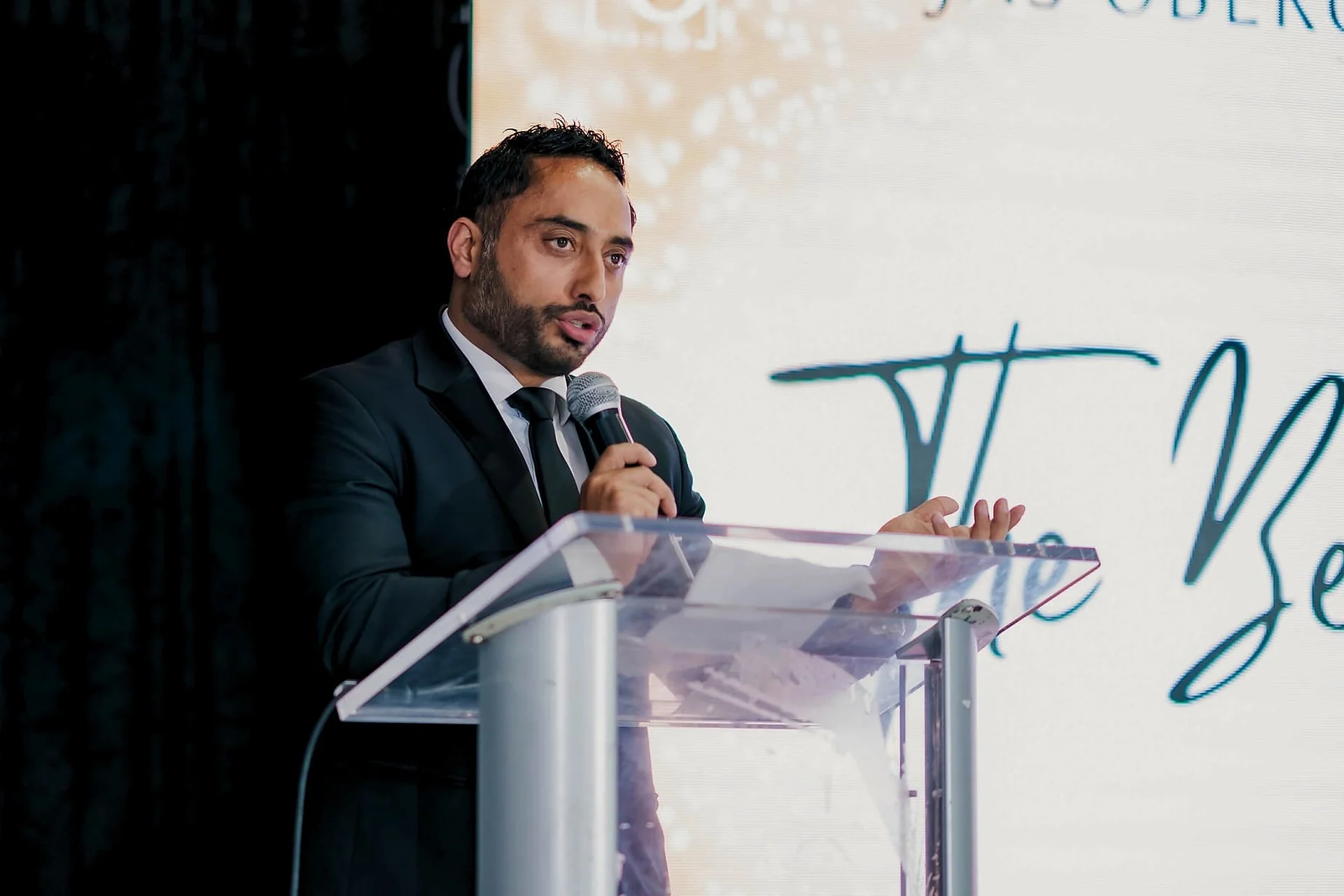 Man in a suit speaking at a podium with script lettering behind, dark curtain left