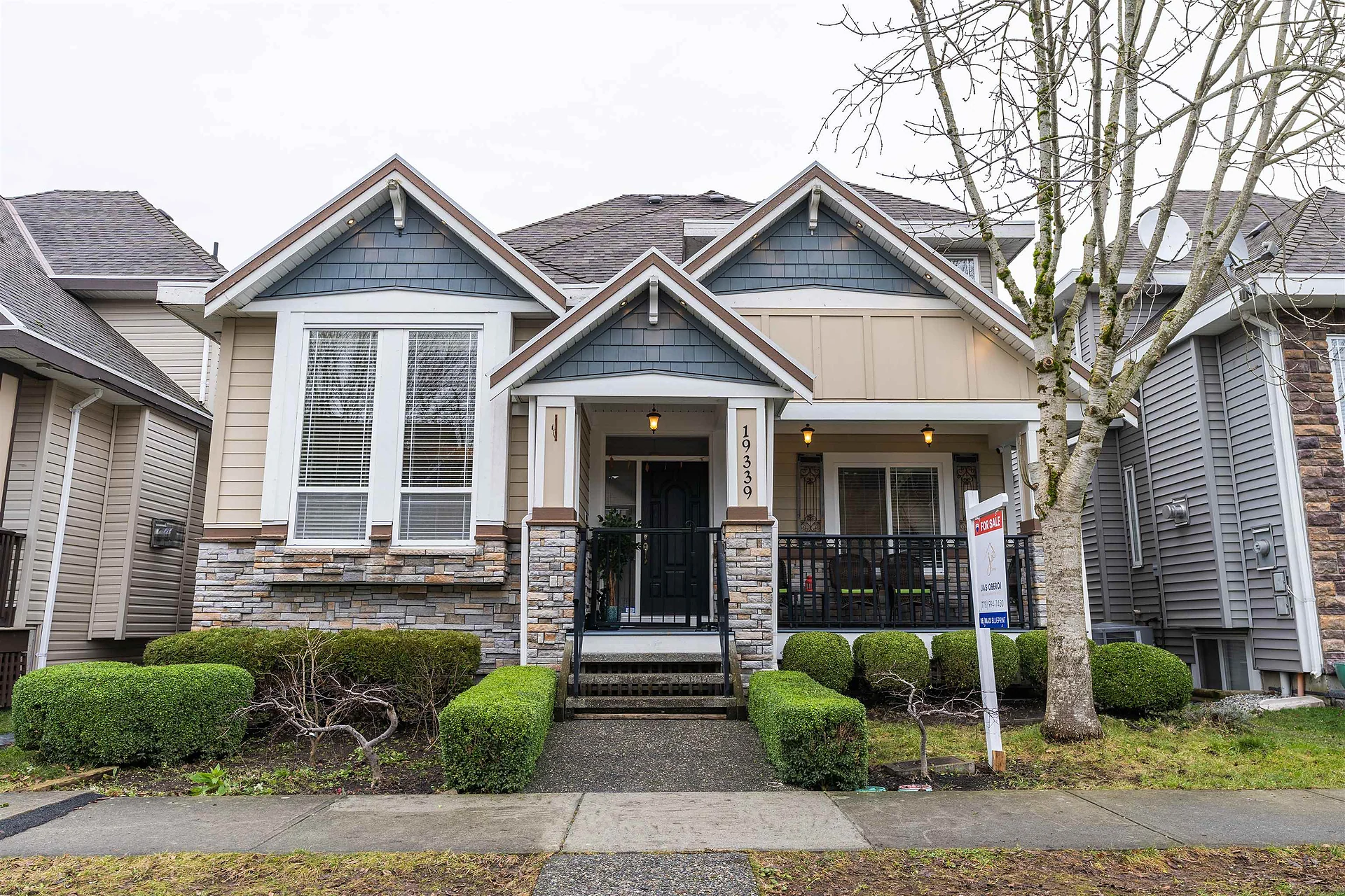 Two-storey craftsman-style house with front porch, hedges, For Sale sign and bare trees
