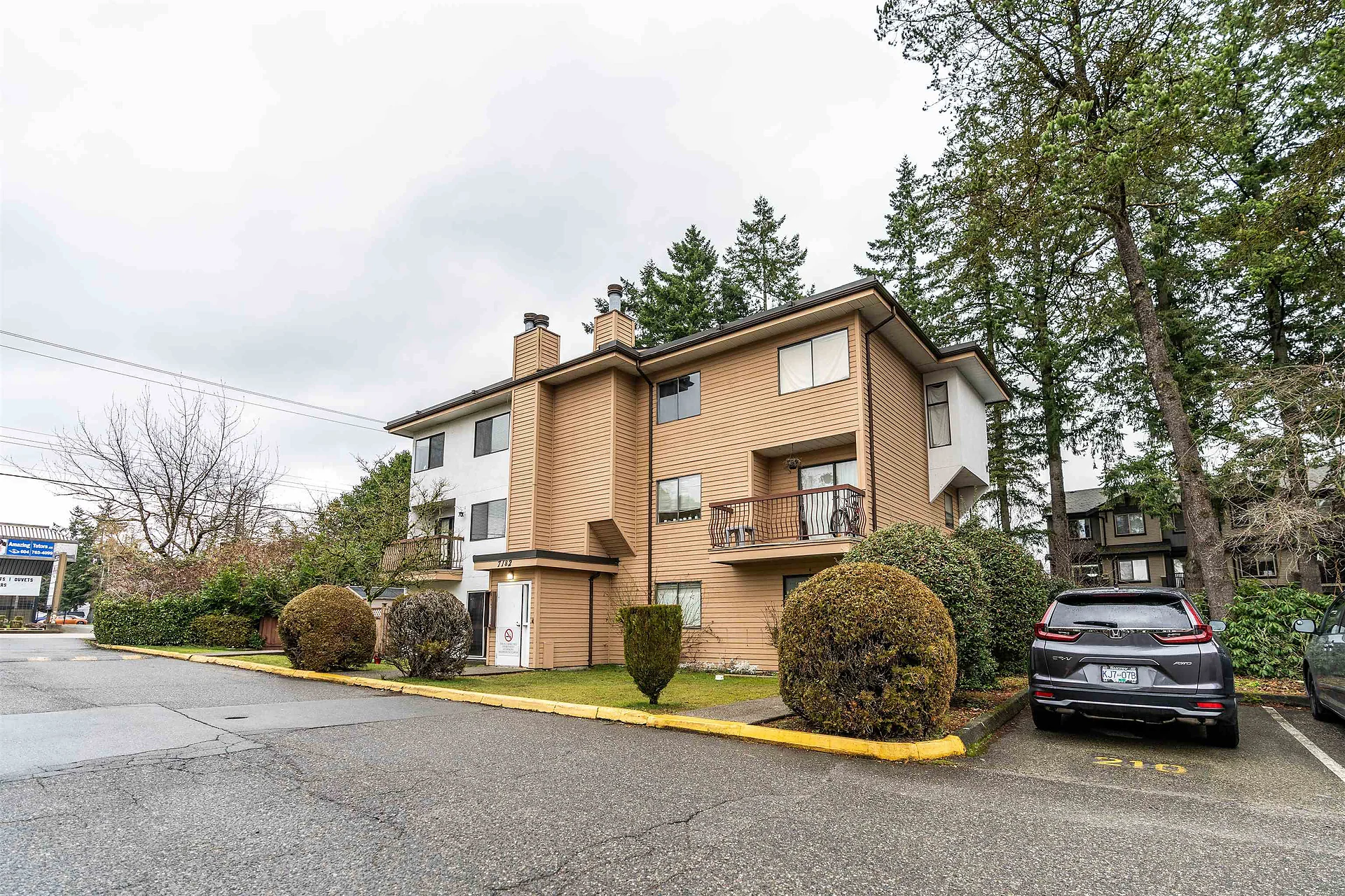 Tan-and-brown three-storey townhouse/condo building beside a parking lot with trees and a parked grey SUV