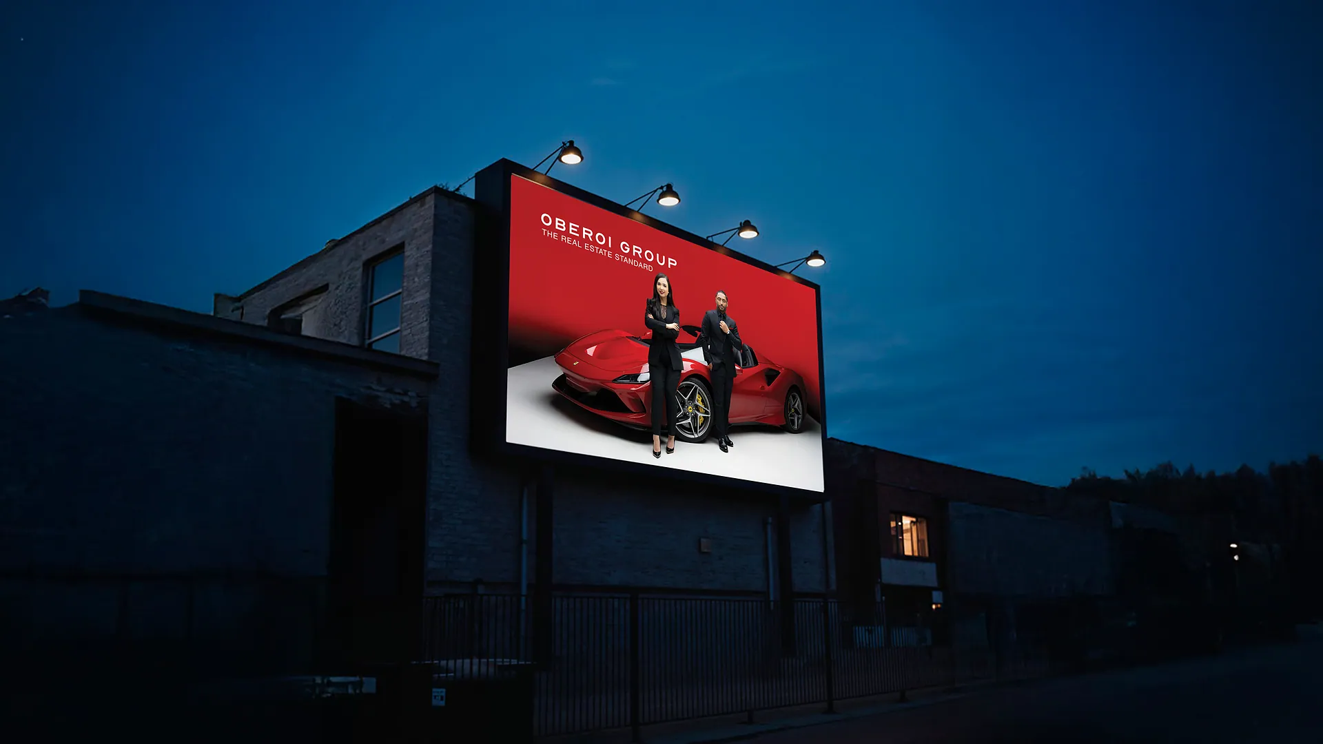 Dusk photo of a lit billboard on a dark building showing two people beside a red sports car with Oberoi Group branding
