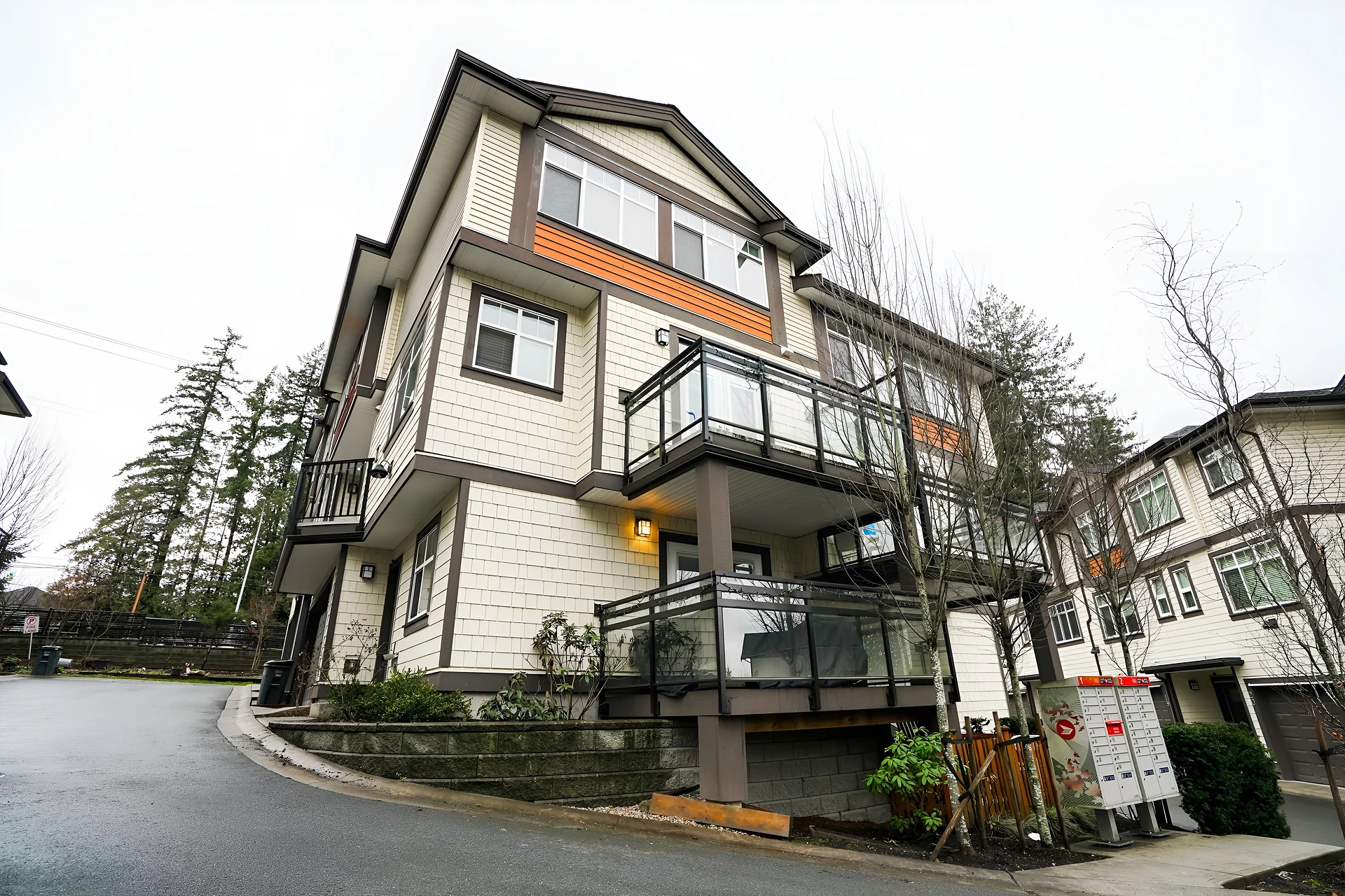 Three-storey townhouse with balconies at a street corner with mailboxes, wet pavement and tall trees