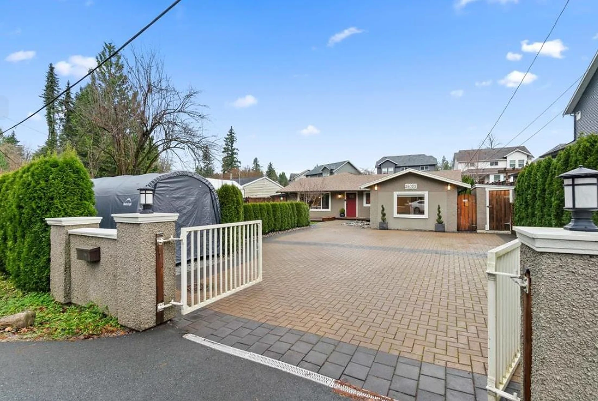 Brick-paved gated driveway with stone pillars leading to a small house, trees behind