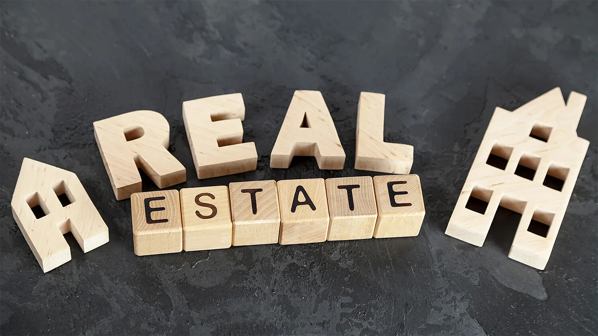 Wooden letter blocks spelling real estate beside small wooden house shapes on dark slate