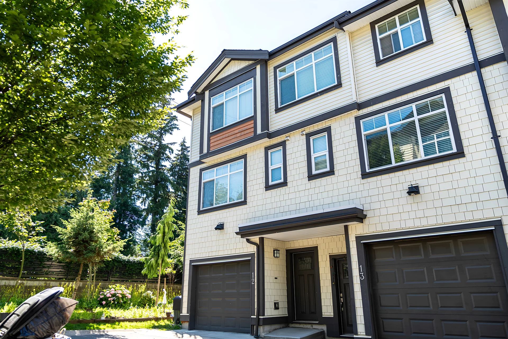 Narrow three-storey townhouse with cream siding and two dark garage doors, trees alongside