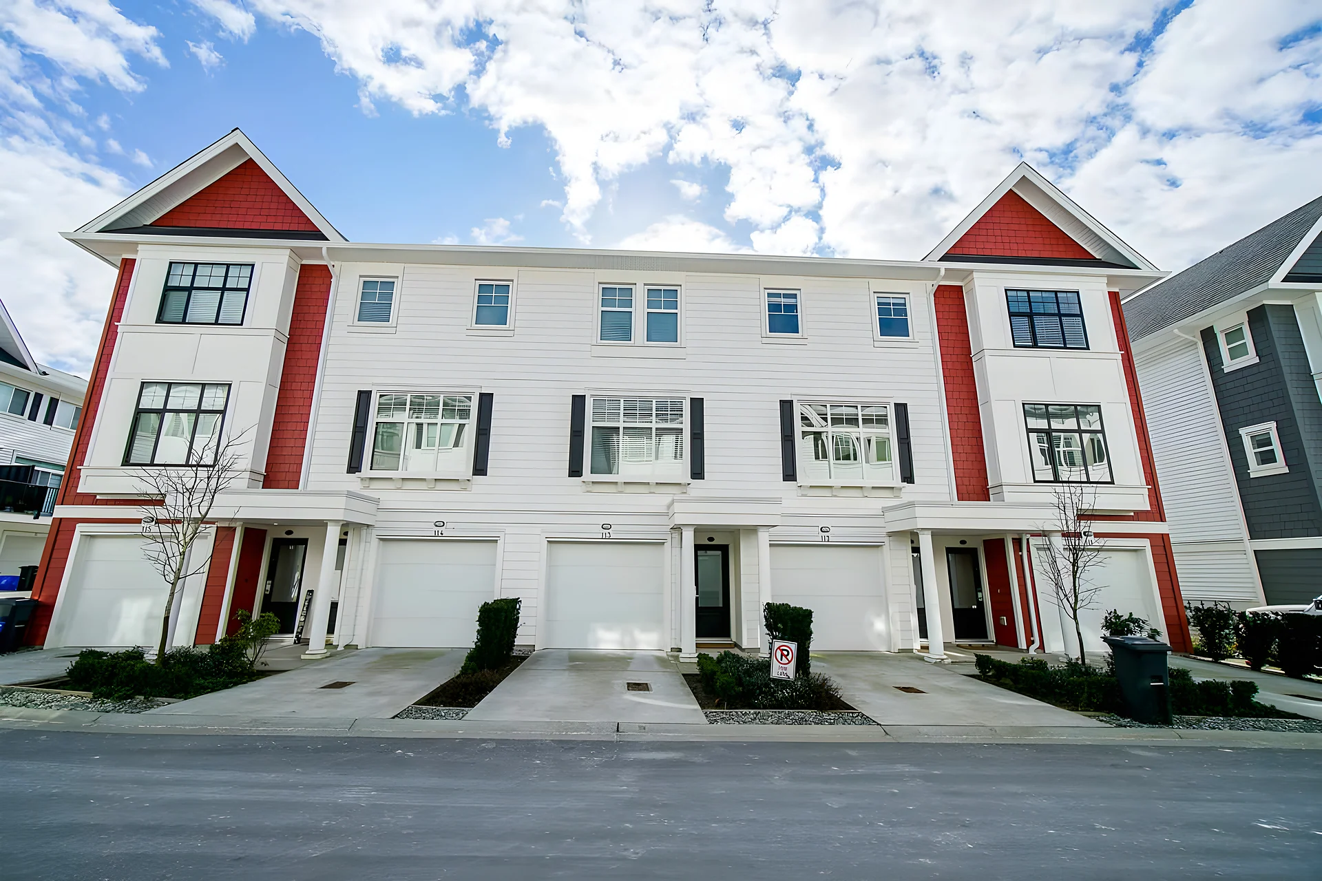 Row of white three-storey townhouses with red accents and ground-level garages on a street under a partly cloudy sky