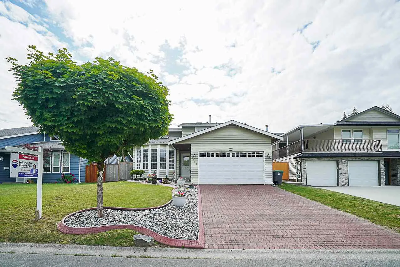 Single-storey house with an attached double garage, brick driveway, a large tree and a real estate sign on the lawn