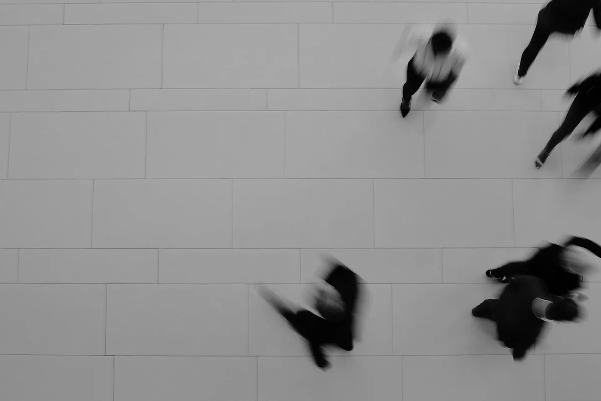 Overhead view of blurred pedestrians in dark clothing walking across large grey paving tiles