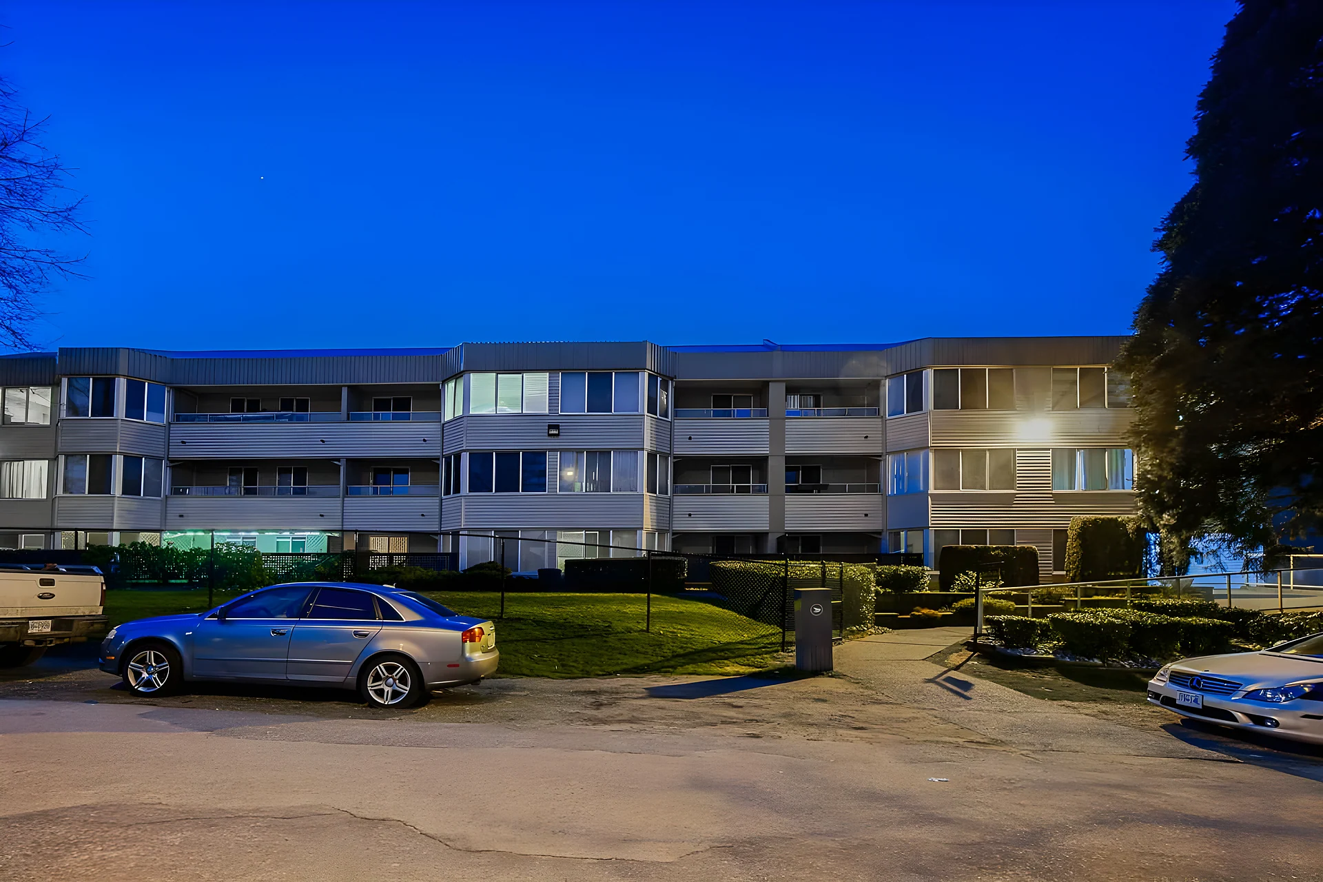 Low-rise apartment building at dusk with lit windows and a car parked in front