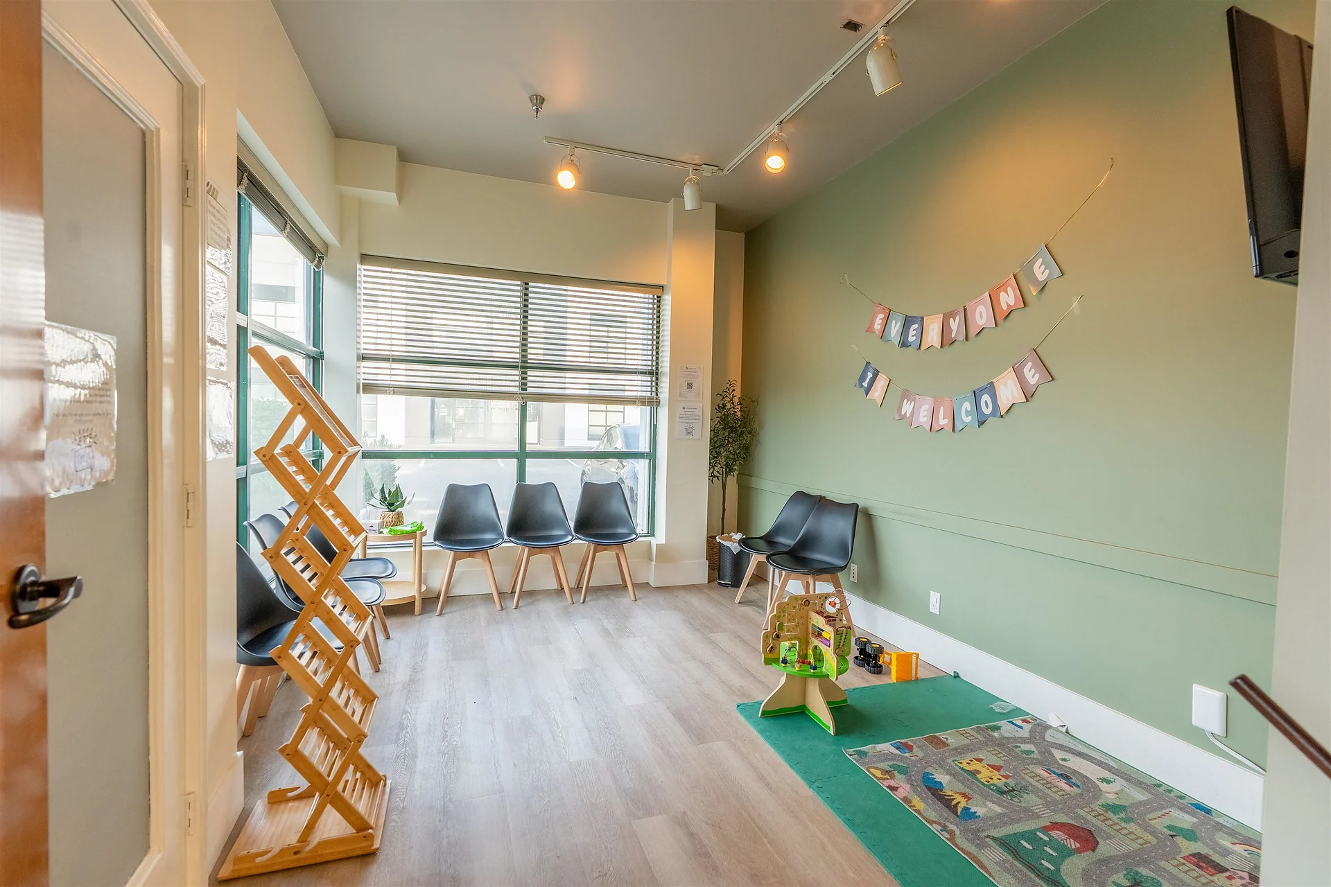 Small room with green wall, wooden shelf, black chairs, play mat and toys, large window