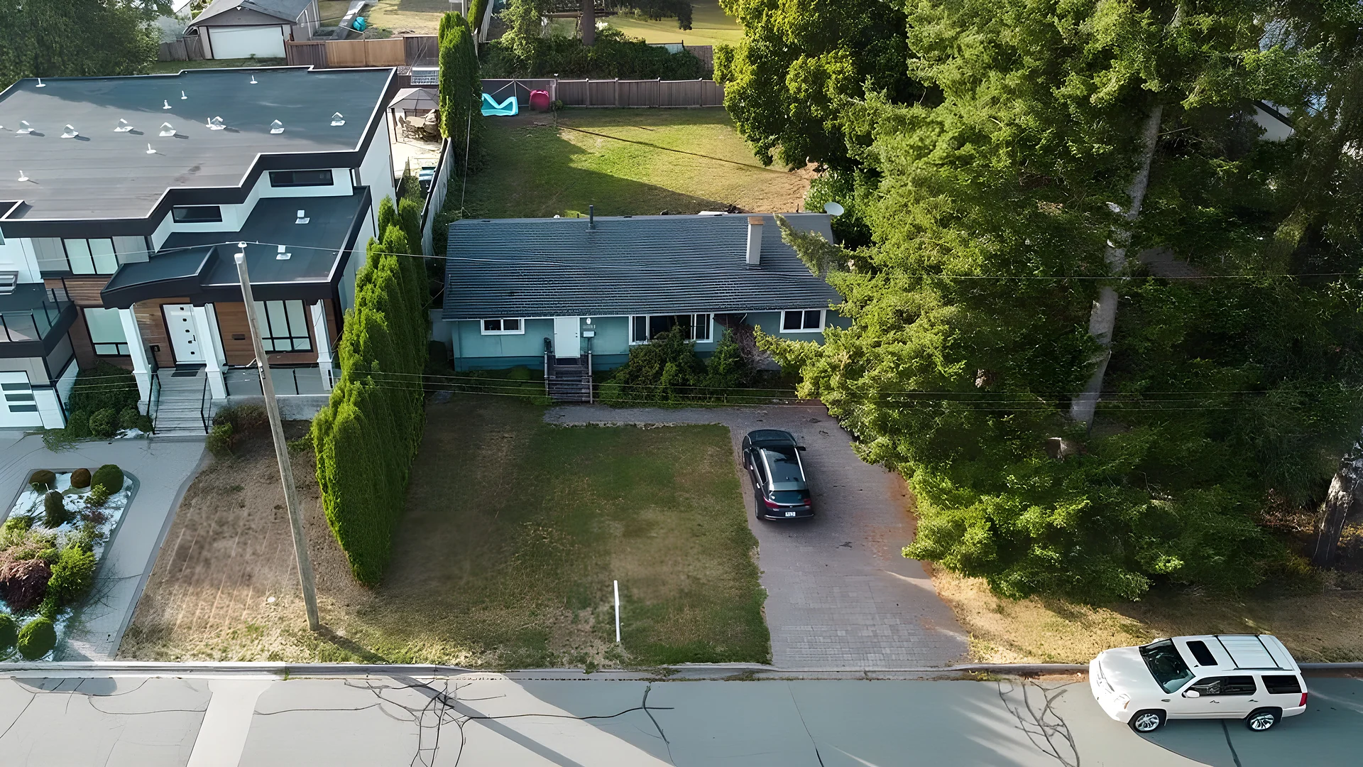 Drone view over a small house with a yard, car in driveway and tall trees; modern houses at left