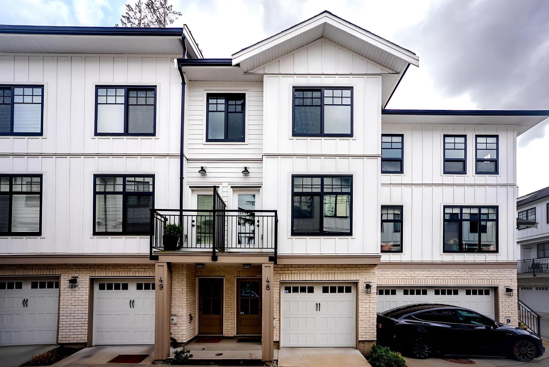 White townhouse building with black window trims, balconies, garages and a black car at right