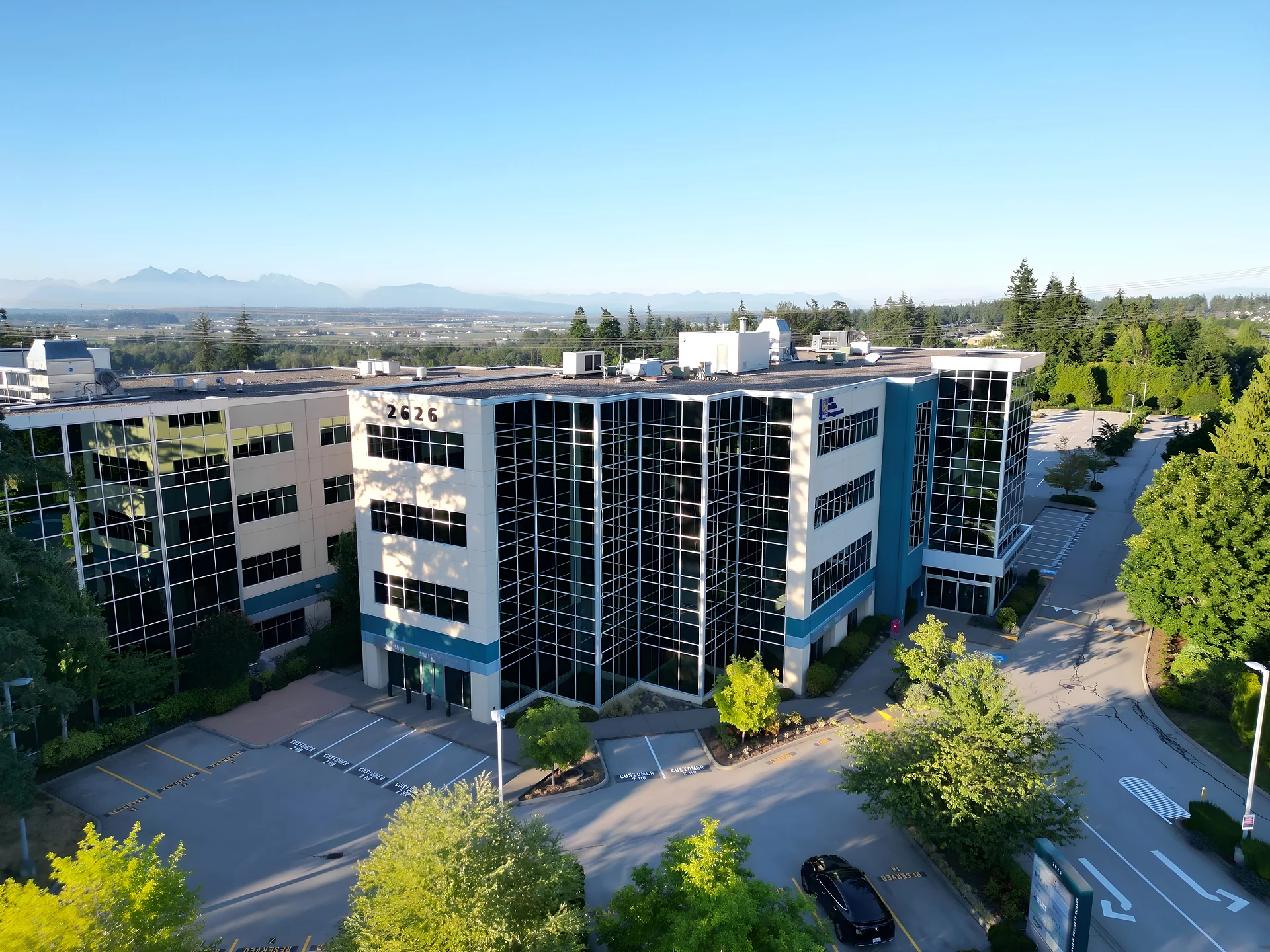 Aerial view of a multi-storey office building with parking lots and trees, mountains in the distance