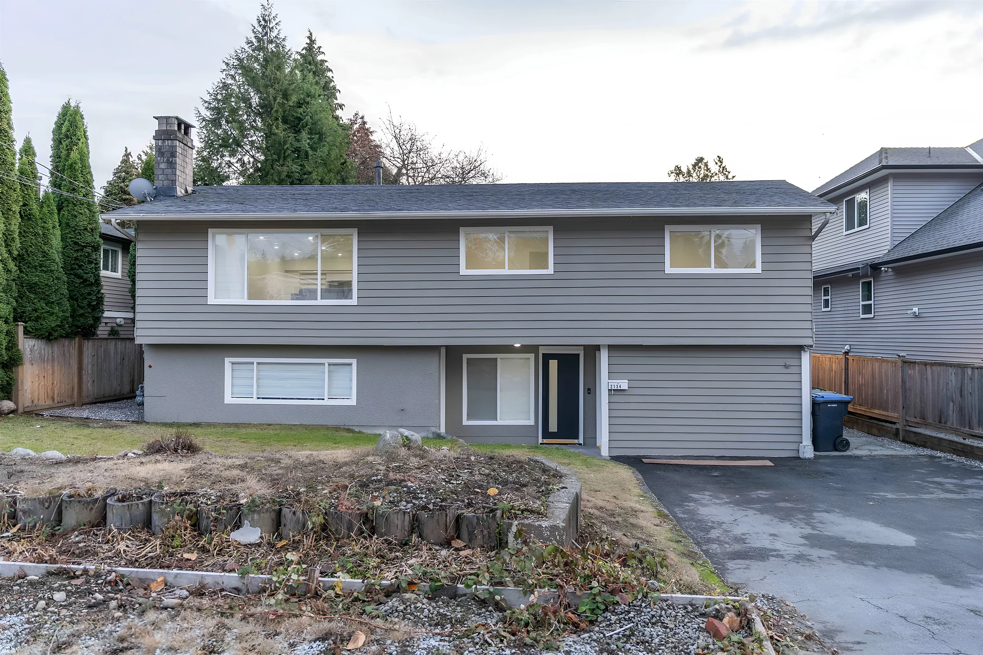 Two-storey grey house with carport and overgrown front yard, fences, overcast