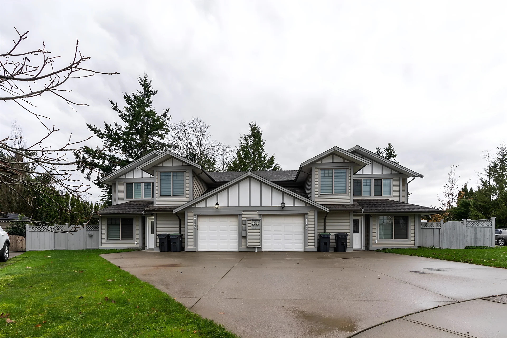 Two-storey duplex with two garage doors, wide driveway and trees behind, overcast