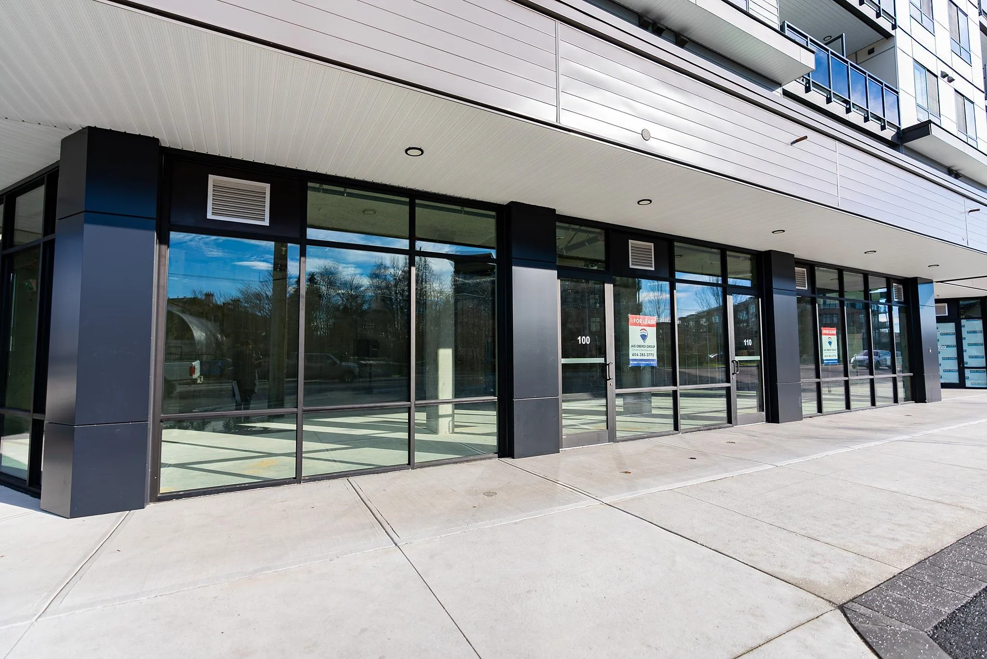 Row of empty glass storefront units at the base of a new building along a sidewalk