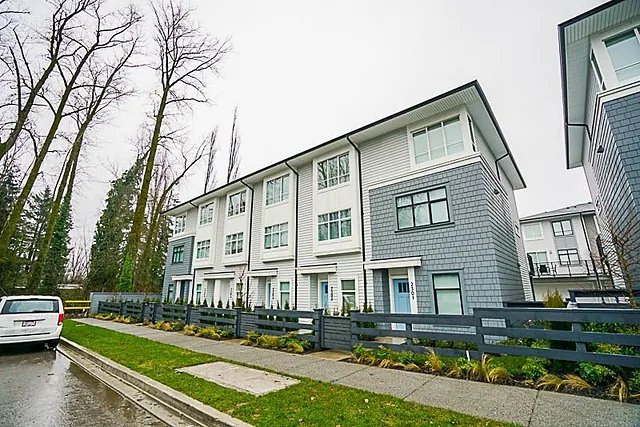 Row of new three-storey townhouses along a sidewalk and a wet street with tall trees behind